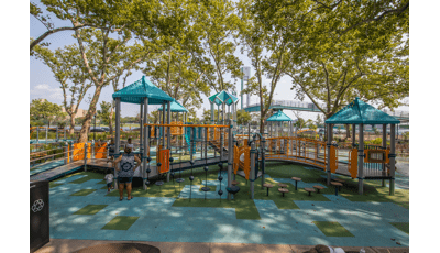 Children and family playing on a playground with light blue roofs.  The playground is shaded by tall green trees. 