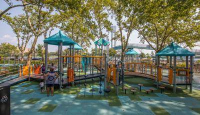 Children and family playing on a playground with light blue roofs.  The playground is shaded by tall green trees. 