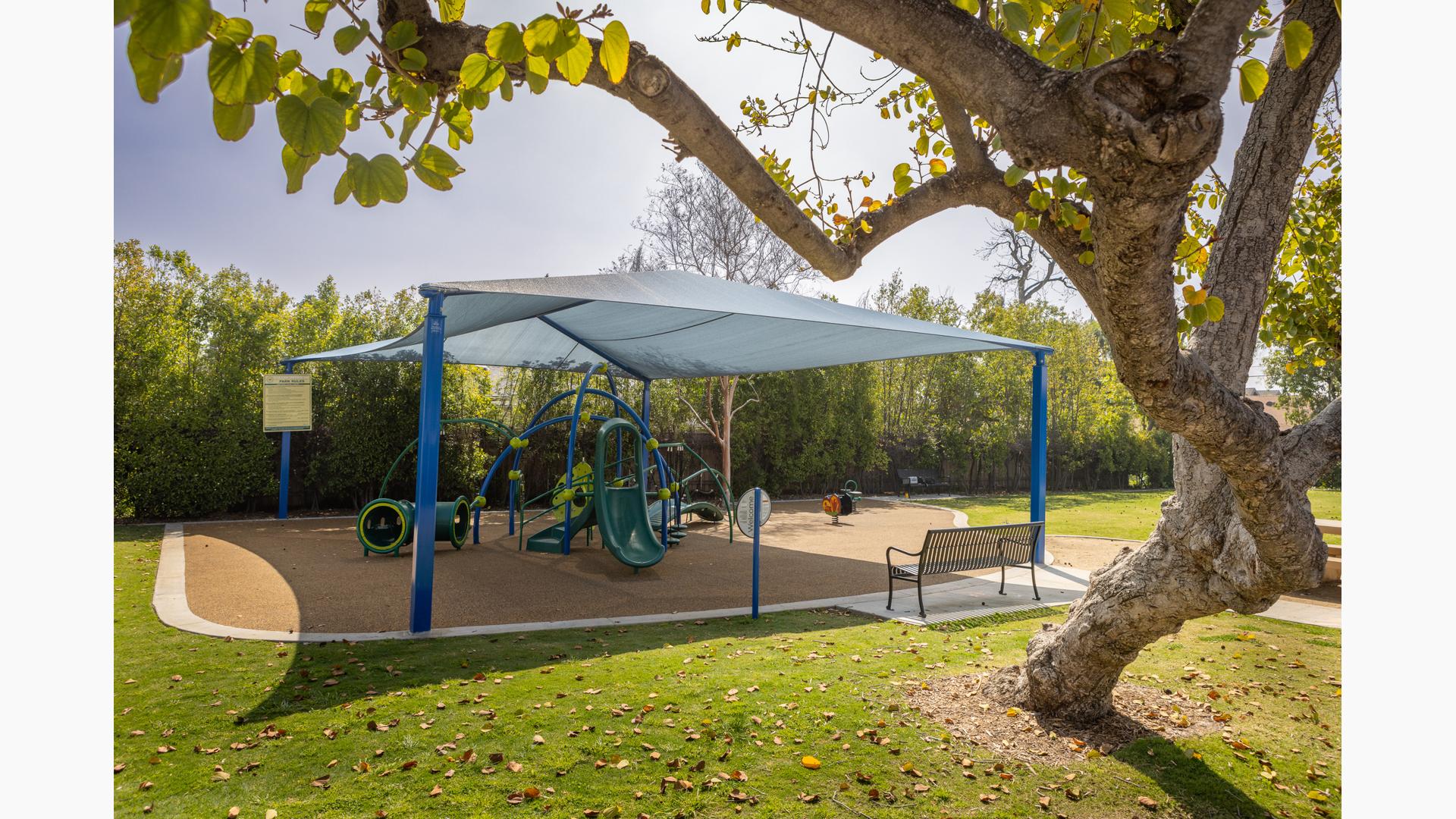 A curvy tree sits off to the side of an outdoor play area with a large shade over a playground  made up of a much of arches and play activities.