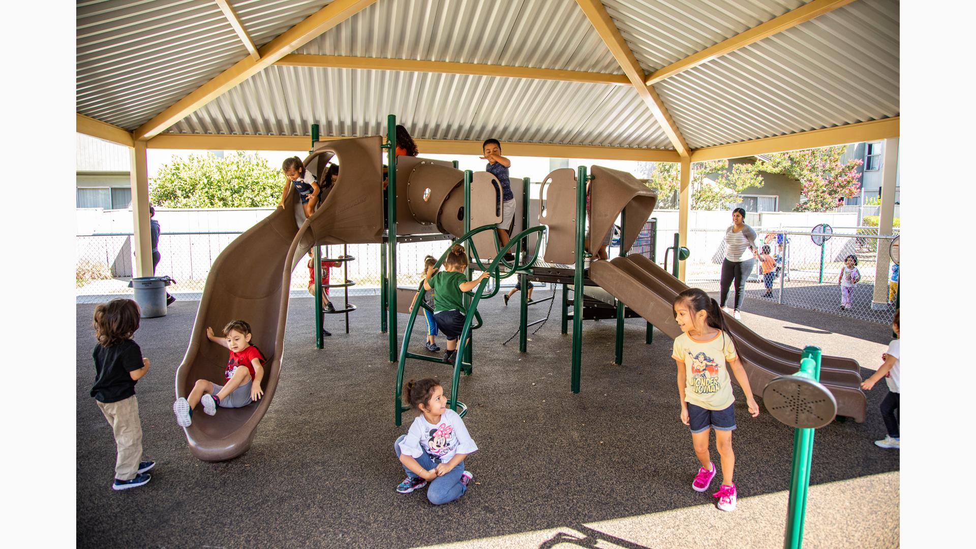 Children playing on a playground structure with climbers and slides with a large tin roof structure overhead.