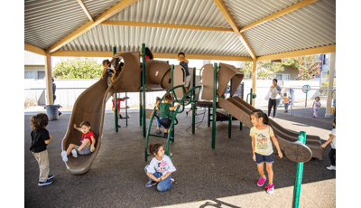 Children playing on a playground structure with climbers and slides with a large tin roof structure overhead.