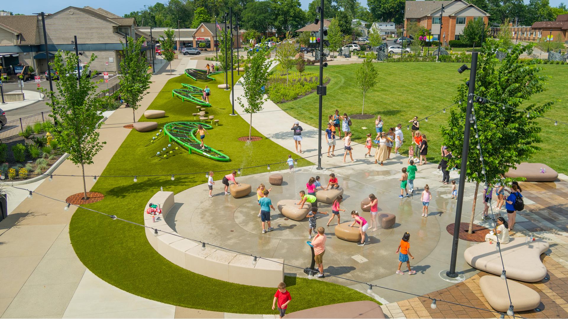 Elevated view of children playing in the water of a small splash pad and three climbing structures lined along the sidewalk or the town square.