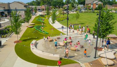 Elevated view of children playing in the water of a small splash pad and three climbing structures lined along the sidewalk or the town square.