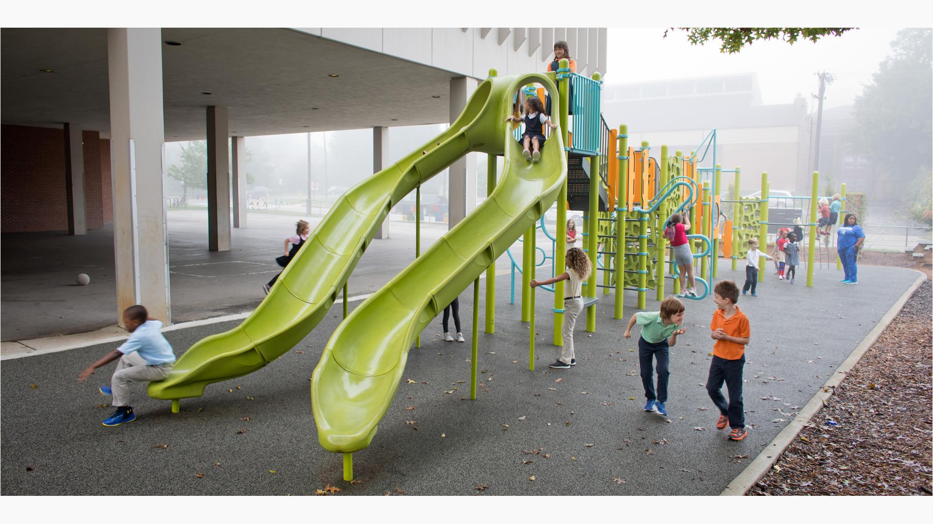 School kids playing in the misting rain on a playground with two limon colored slides in the foreground. 