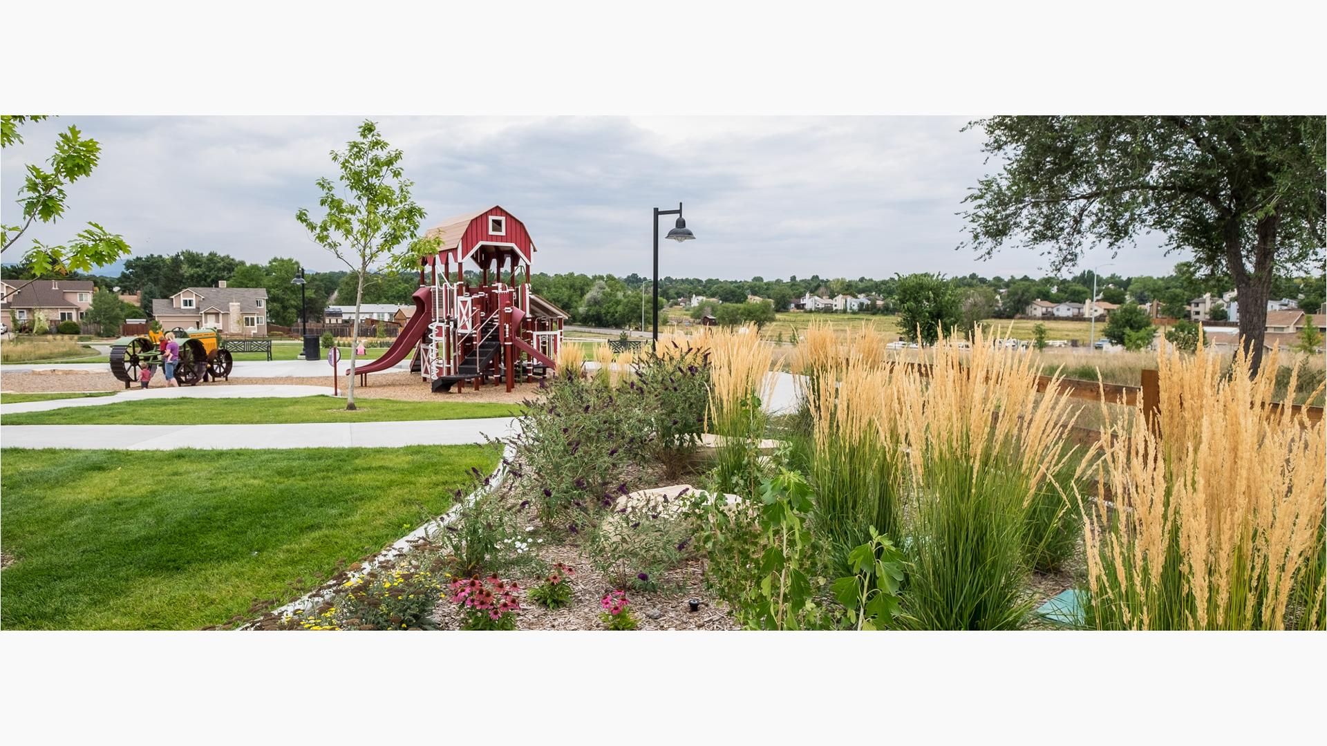 Children play on a full sized tractor play peice next to a barn themed playstructure with tall grasses and flowers in a garden in the forground. 