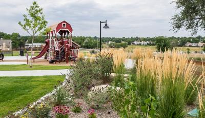 Children play on a full sized tractor play peice next to a barn themed playstructure with tall grasses and flowers in a garden in the forground. 