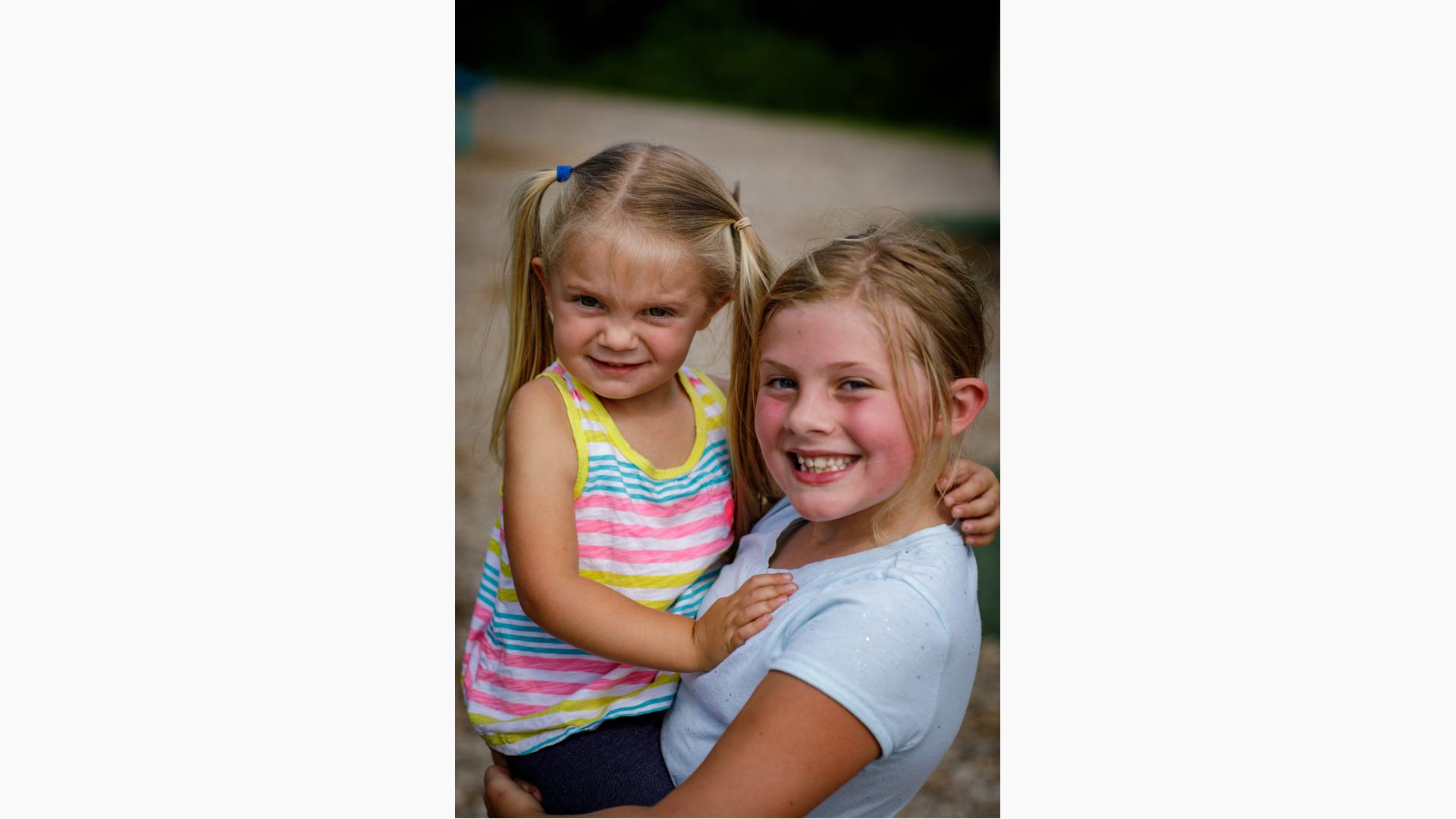 Girl in white shirt smiles as she holds little girl in striped shirt.