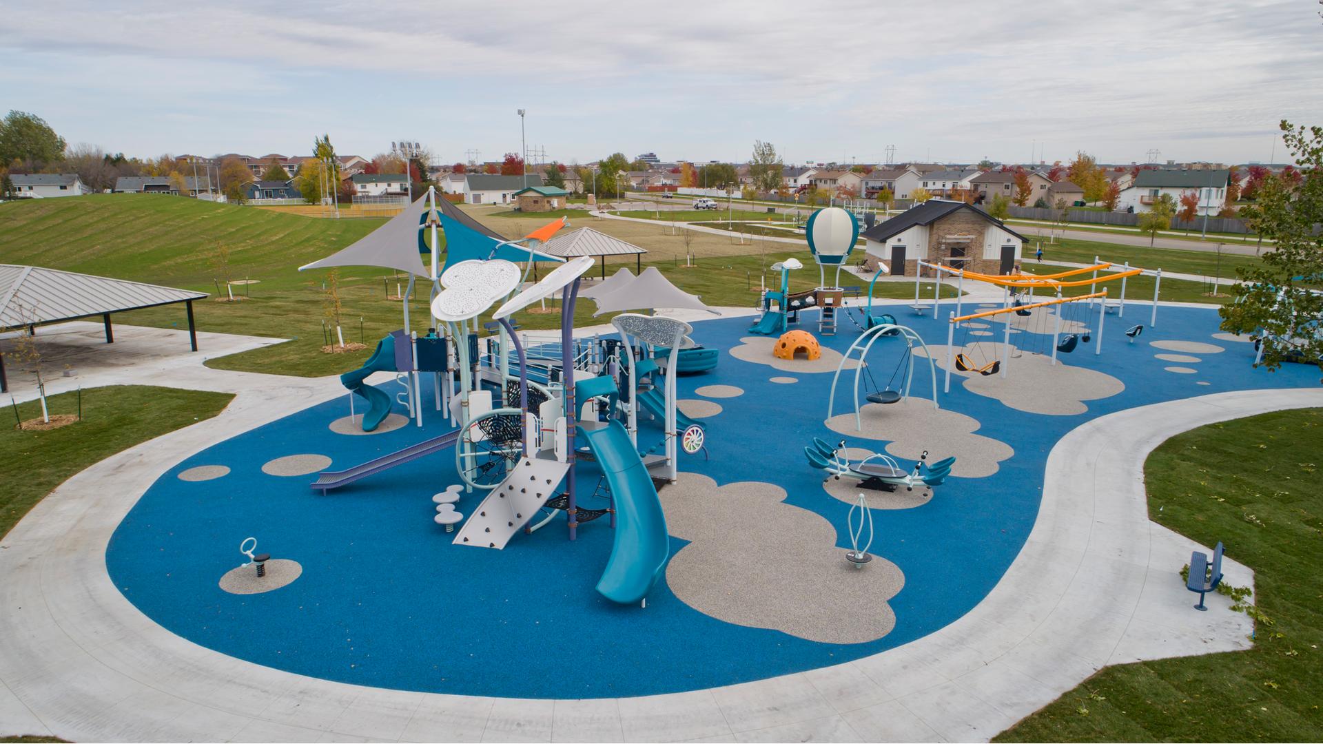 Large cloud and hot air balloon-themed play structures for all ages is surrounded by spinners, climbers, and swing set all set on a safety surfacing designed with grey cloud shapes. 