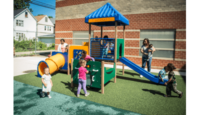 Children play all over a toddler accessible play structure with play panels, slides, and climbers all next to a brick building.