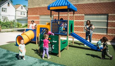 Children play all over a toddler accessible play structure with play panels, slides, and climbers all next to a brick building.