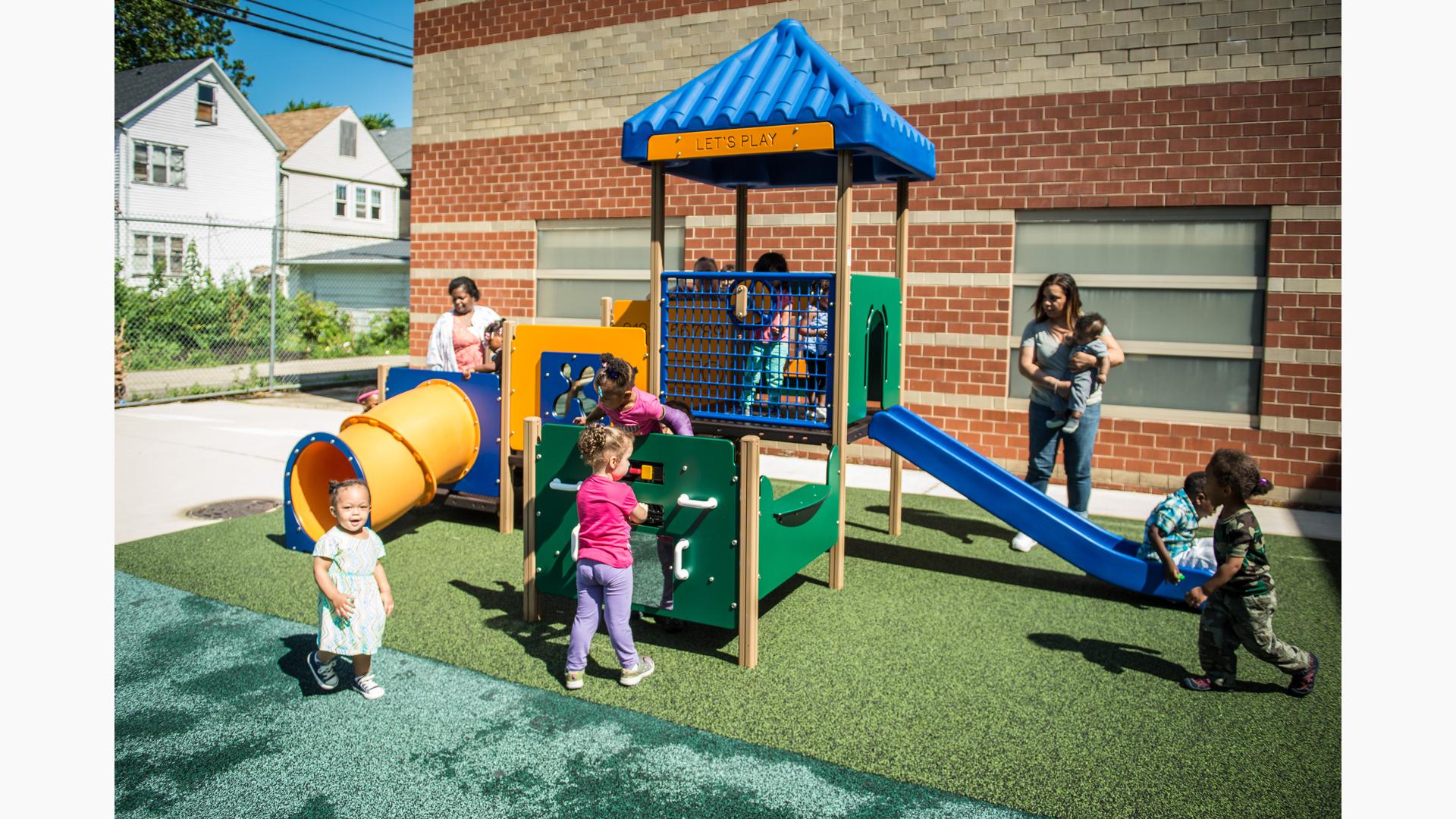 Children play all over a toddler accessible play structure with play panels, slides, and climbers all next to a brick building.