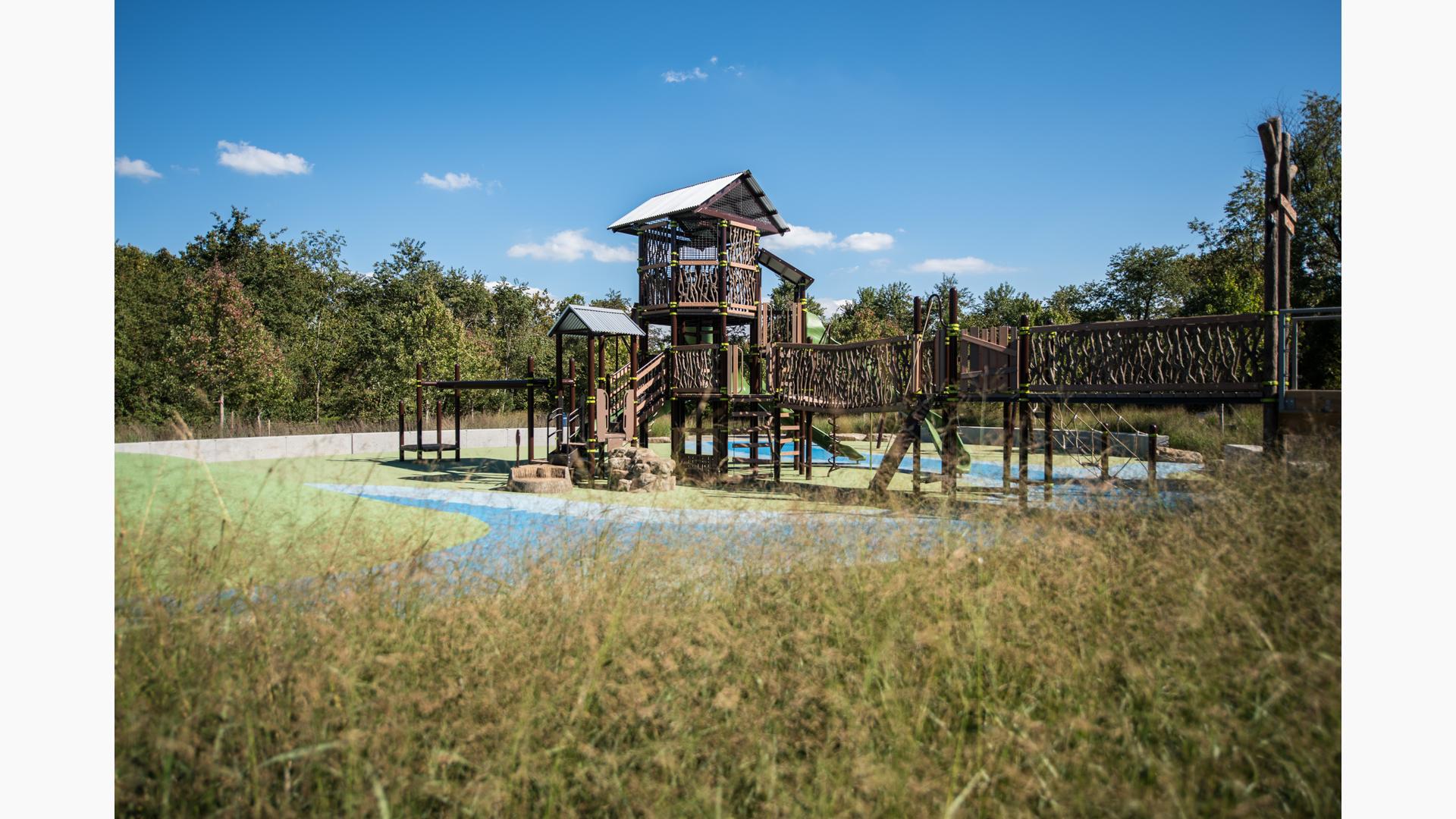 Nature-inspired playground with look-a-like log bridge. Playground is a two-story tower with a metal steel roof. 