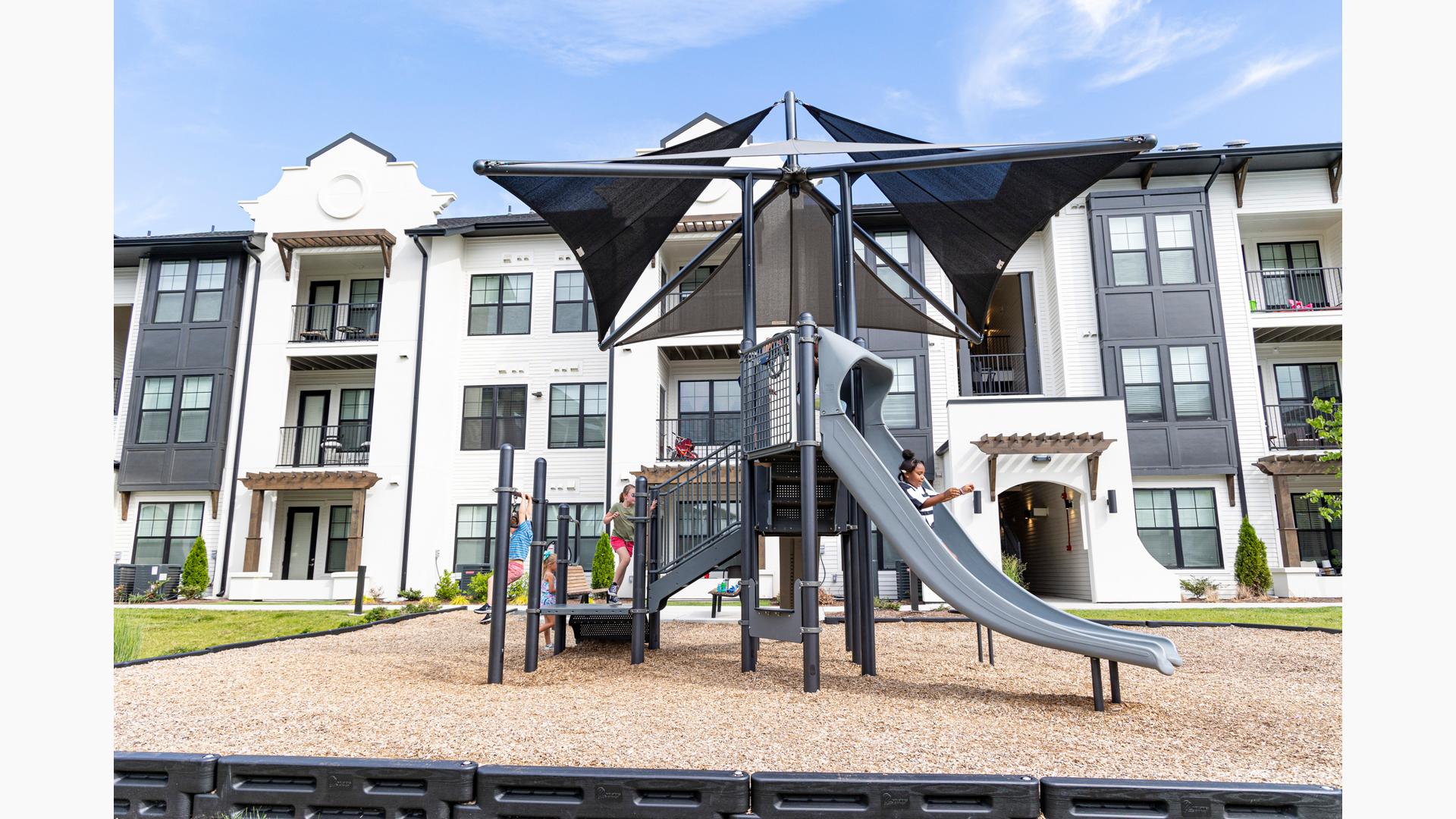 Children play on a playground structure with climbers slides, two tear decking, and shade system over head that sits next to an apartment building.
