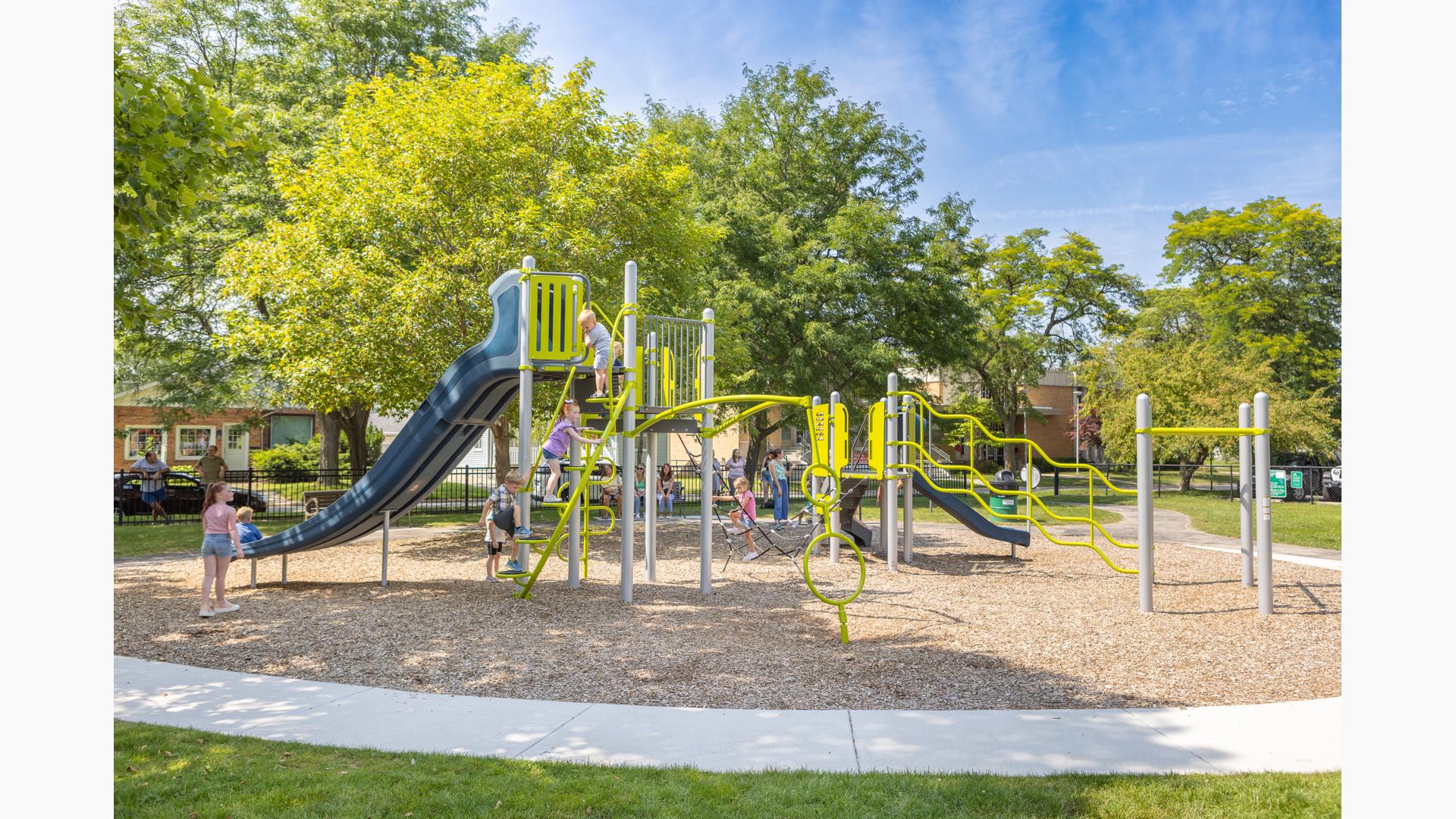 Children play on a play structure at a community park surrounded by lush green trees on a sunny day.