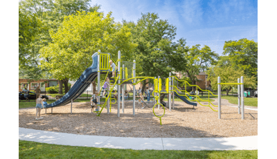 Children play on a play structure at a community park surrounded by lush green trees on a sunny day.