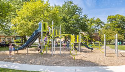 Children play on a play structure at a community park surrounded by lush green trees on a sunny day.