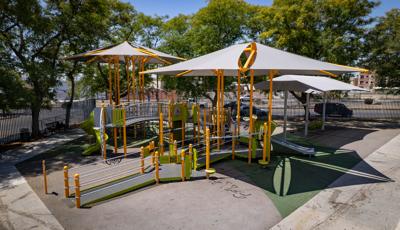 Slightly elevated view of a fenced in large inclusive playground with accessible ramps, bridges, play panels, and two large overhead shade systems. Two bays of swings sit just behind the play structure with its own large shade system.