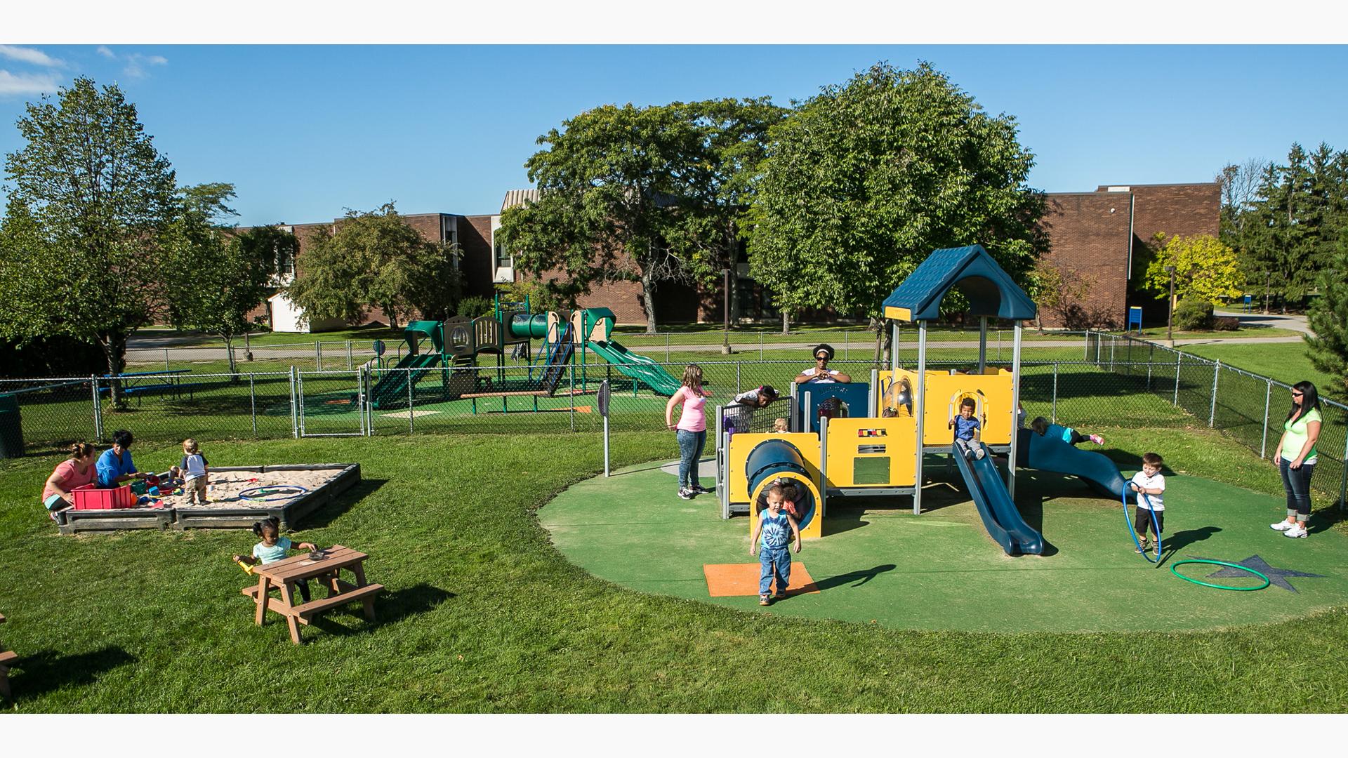 Mothers stand near by as children play on a toddler accessible playground and sand box all surrounded by a chain-link fence. 