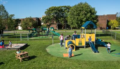 Mothers stand near by as children play on a toddler accessible playground and sand box all surrounded by a chain-link fence. 