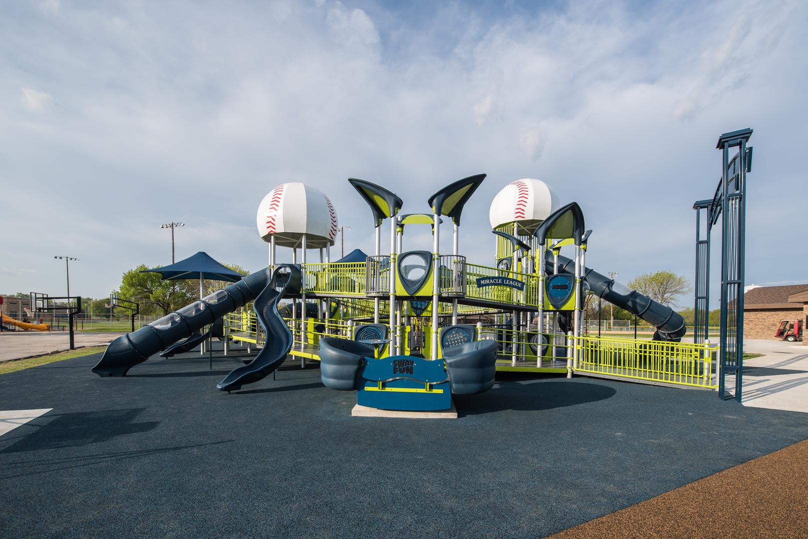 Green, blue and silver playground with 2 baseball dome roofs. Ramps and tall blue slides. Basketball courts in background.