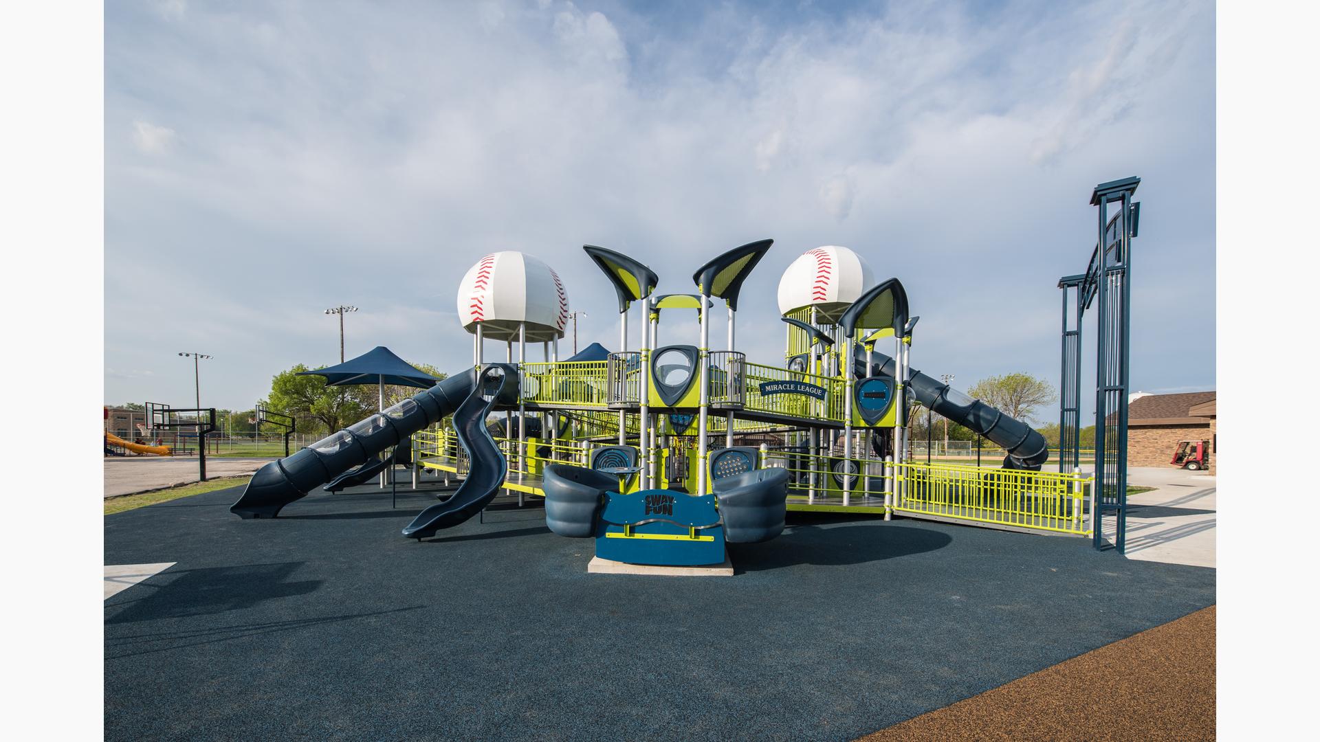 Green, blue and silver playground with 2 baseball dome roofs. Ramps and tall blue slides. Basketball courts in background.