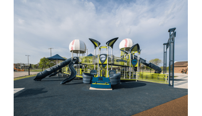 Green, blue and silver playground with 2 baseball dome roofs. Ramps and tall blue slides. Basketball courts in background.