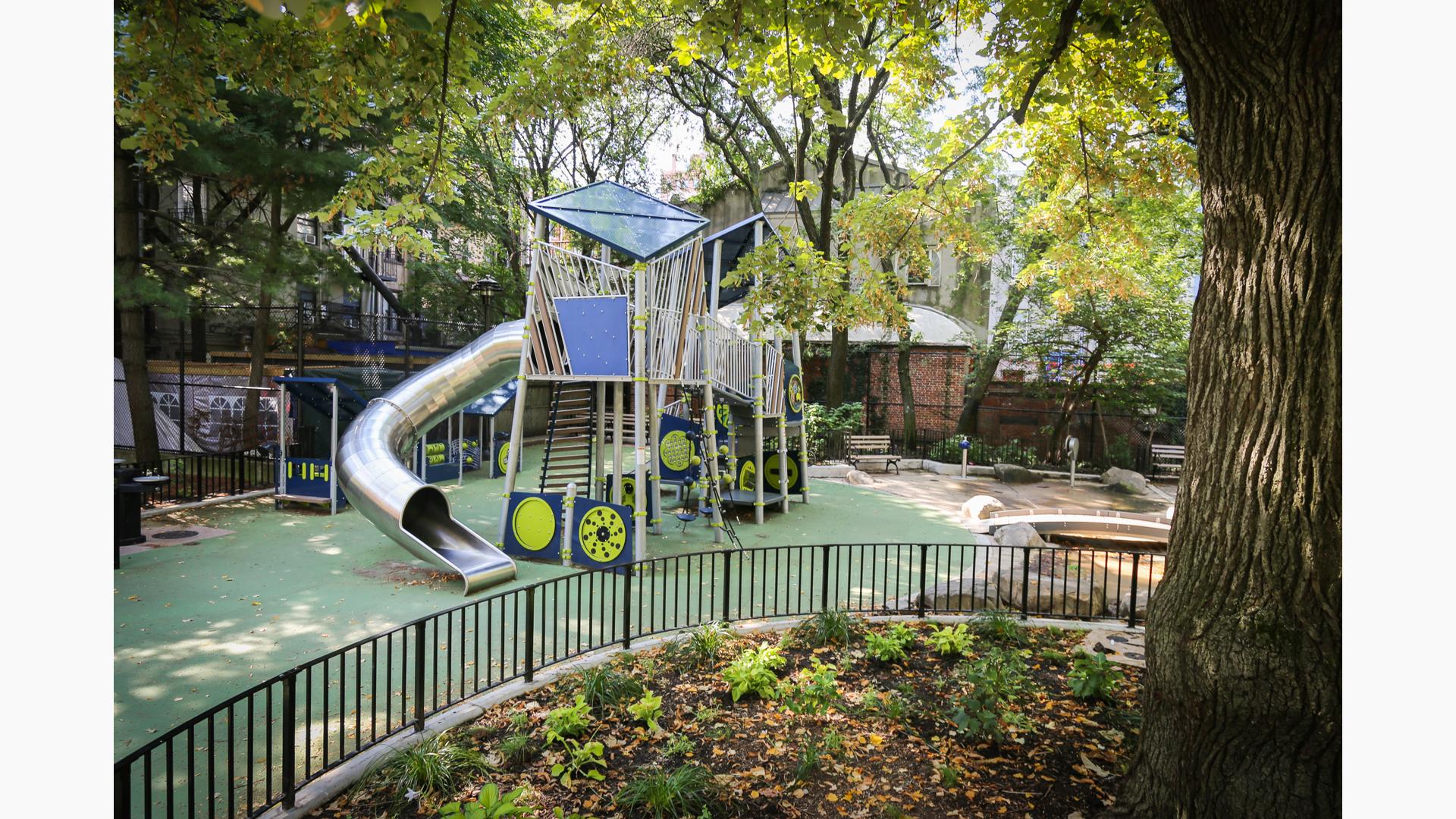 View past a large oak tree of a custom modern designed play structure with large stainless steel tubular slide. 