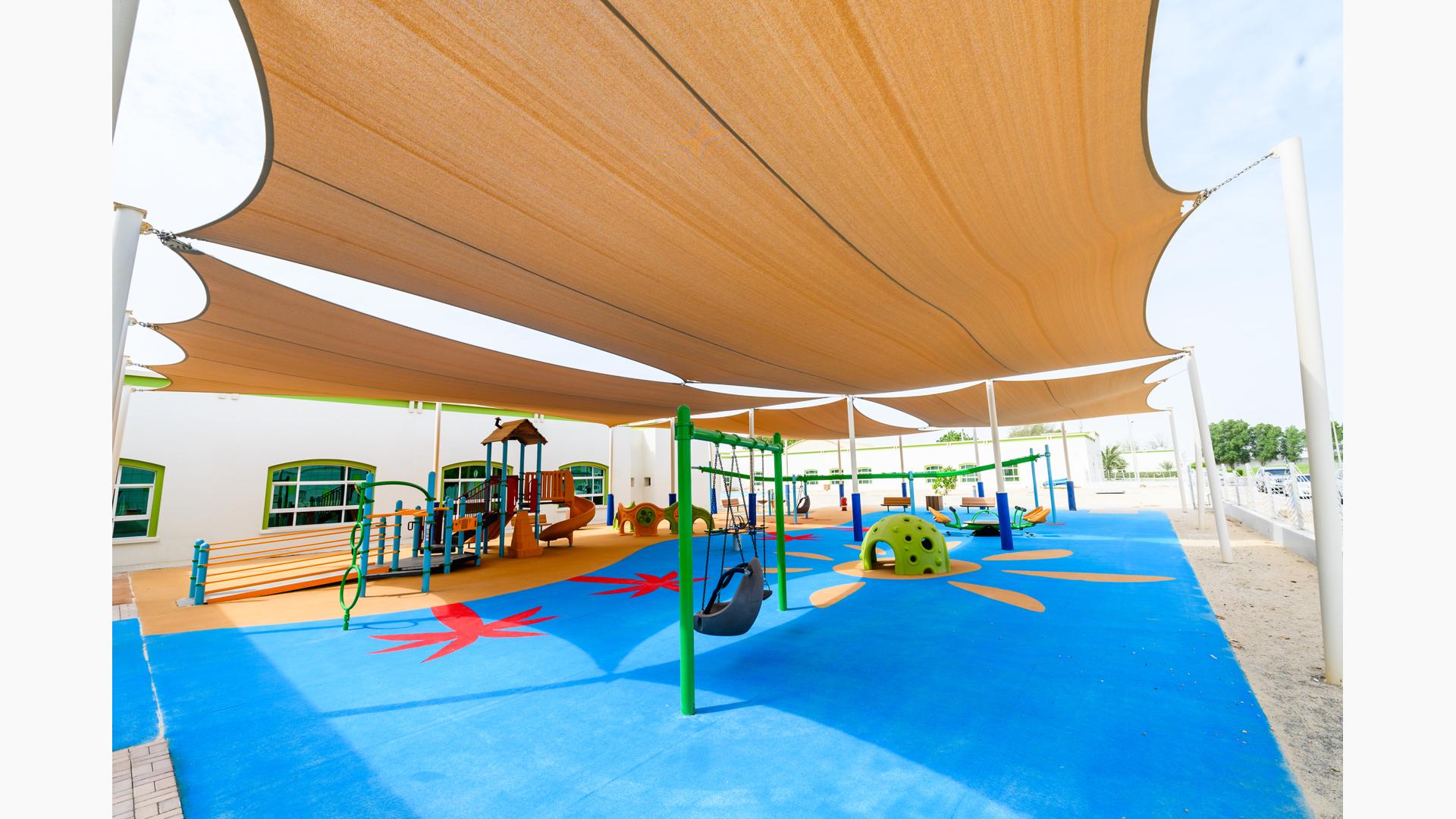 Large tan shade cover this play area with many inclusive playground elements including a ramped playground structure, single post swing, quiet area for kids to take a break and an accessible teeter totter all over colorful unitary surfacing.