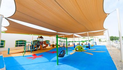 Large tan shade cover this play area with many inclusive playground elements including a ramped playground structure, single post swing, quiet area for kids to take a break and an accessible teeter totter all over colorful unitary surfacing.