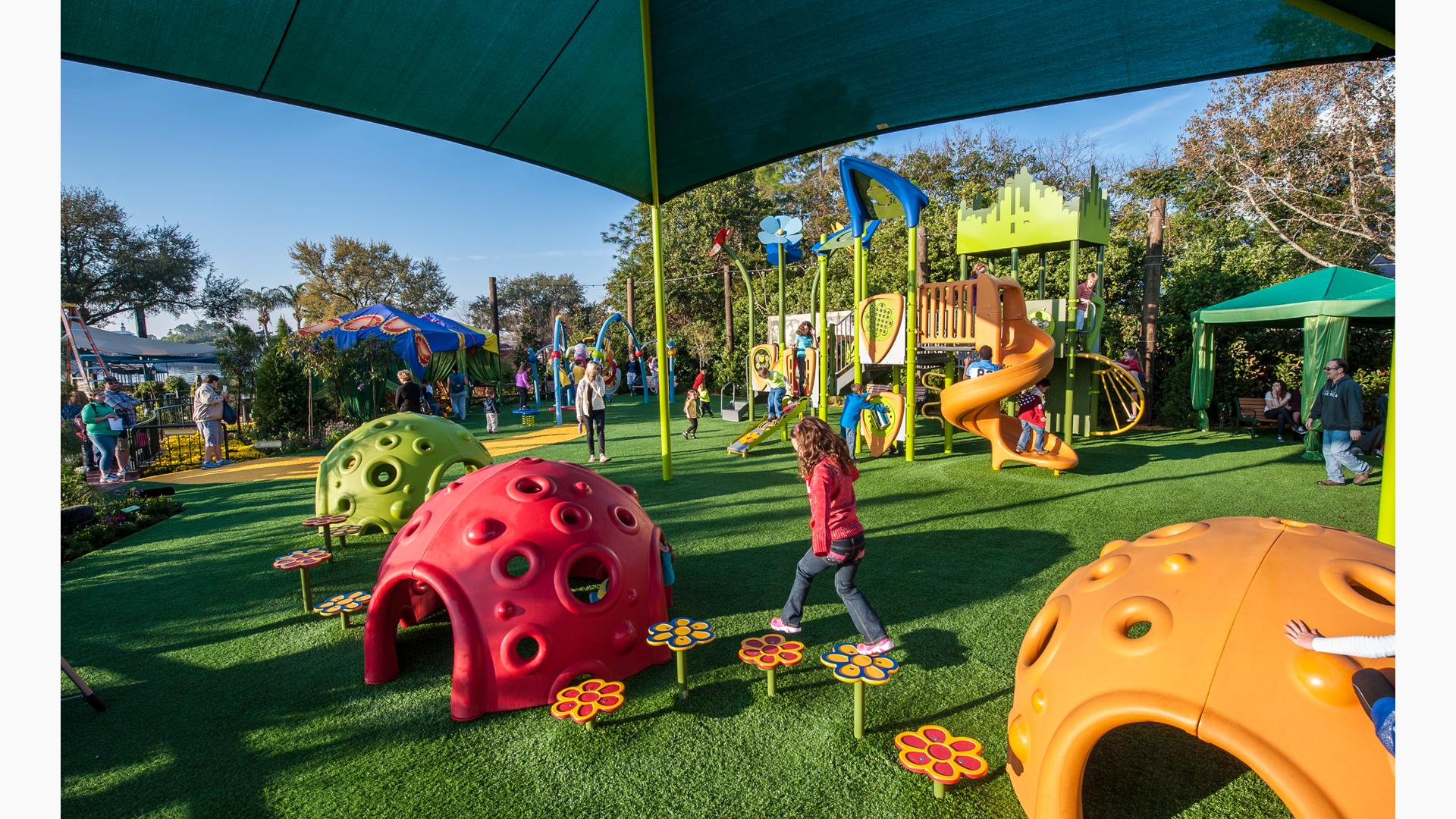 Girl in red jacket walks across flower pod toppers siting between two Cozy domes. A SkyWays shade canopy covers the area. Other children play on the Wizard of Oz-inspired play structure in the background.