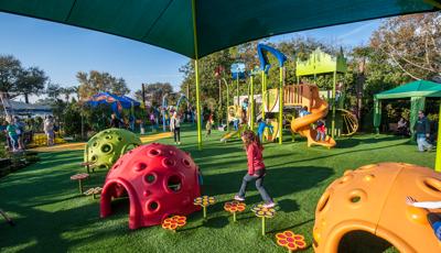 Girl in red jacket walks across flower pod toppers siting between two Cozy domes. A SkyWays shade canopy covers the area. Other children play on the Wizard of Oz-inspired play structure in the background.