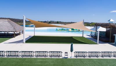Elevated view of a large crystal blue pond in the background with a massive square tan shade over a square sitting space with artificial grass and tiki hut on one side and a building on the other.