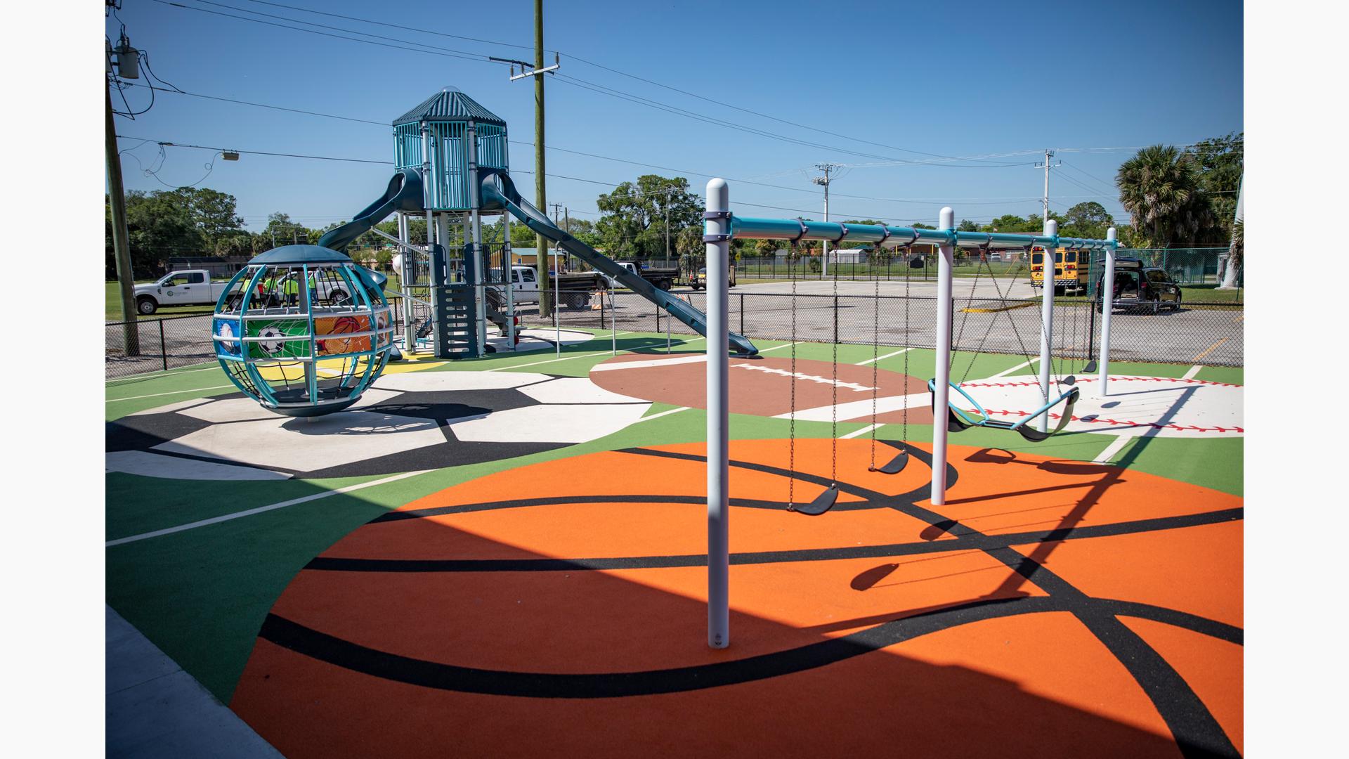 A fenced in play area with sports themed safety surfacing has several bays of swings, a large globe shaped multi-person spinner and a two story playground tower with climbers a slide.