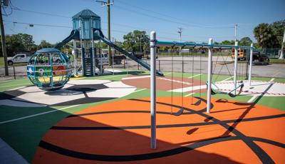 A fenced in play area with sports themed safety surfacing has several bays of swings, a large globe shaped multi-person spinner and a two story playground tower with climbers a slide.