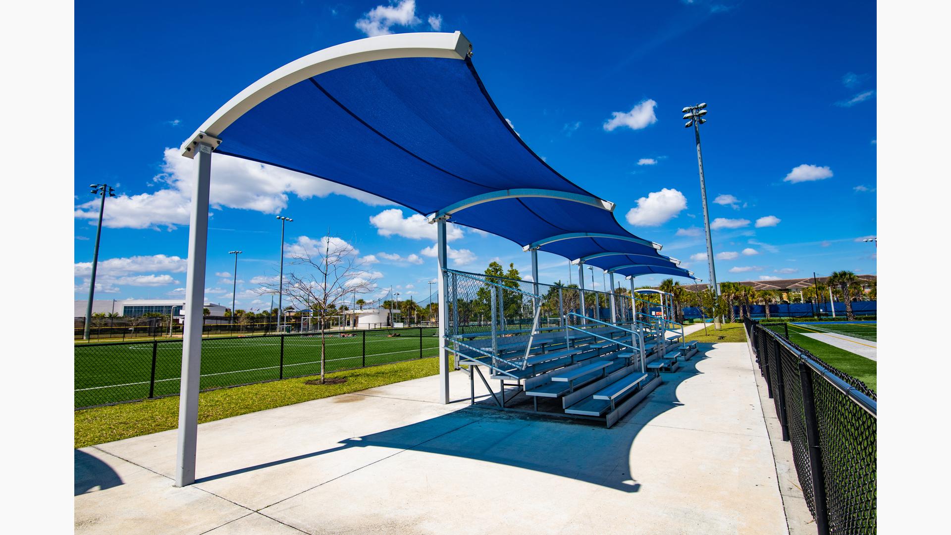 Blue curved shade located at an athletic field providing shade over bleachers. 