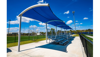 Blue curved shade located at an athletic field providing shade over bleachers. 