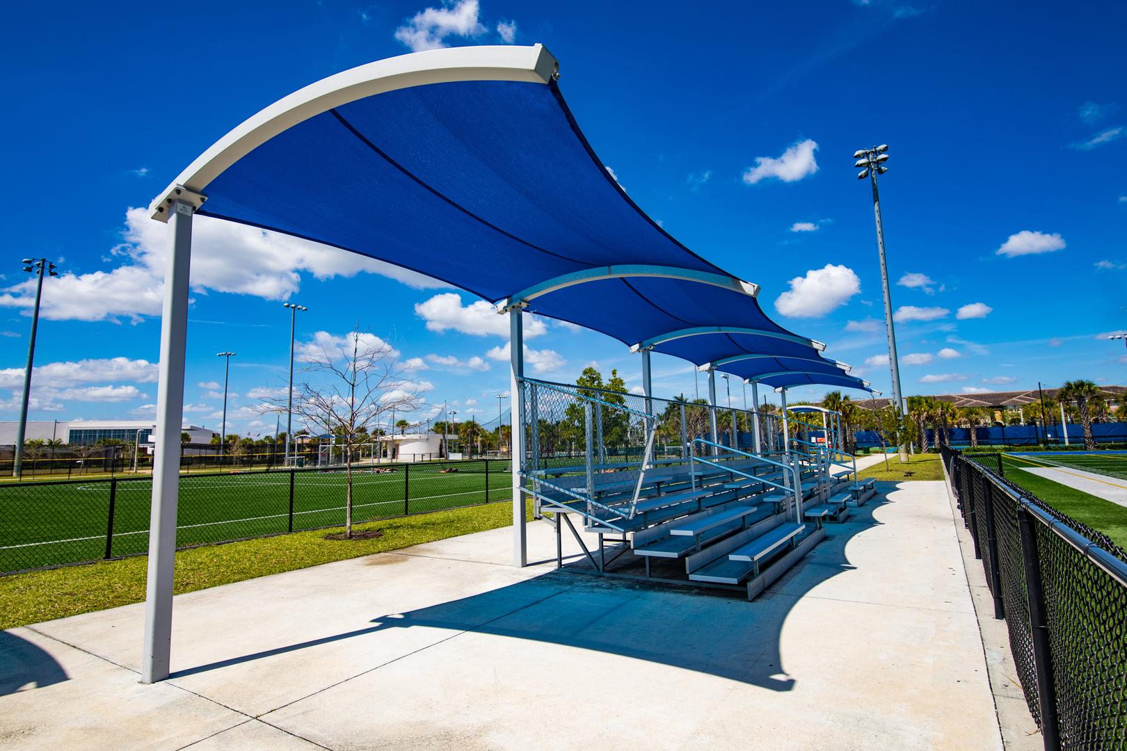 Blue curved shade located at an athletic field providing shade over bleachers. 