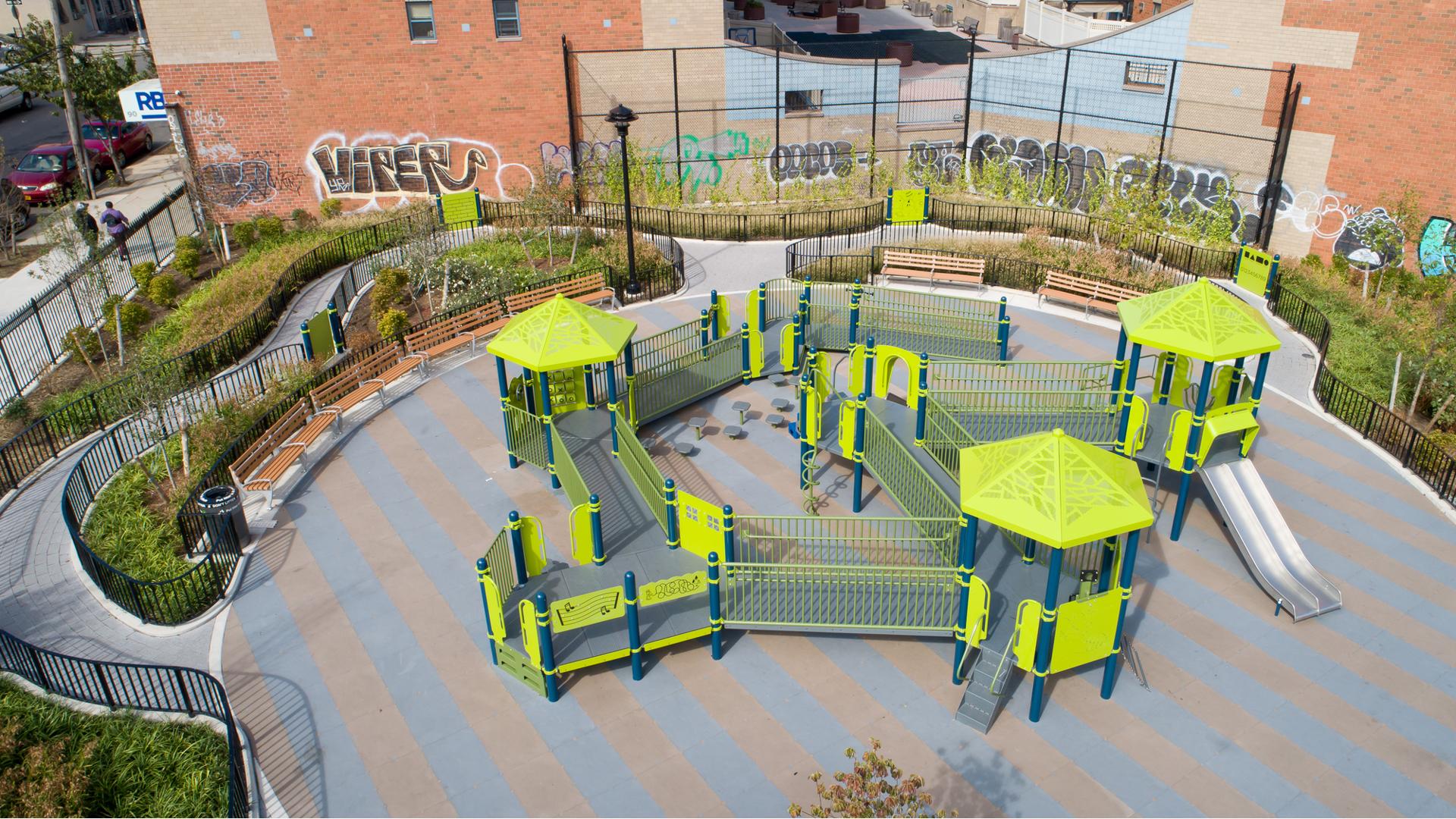 Full elevated view of an inclusive playground with accessible ramps and play panels set next to a brick building with a mural painted on the side.