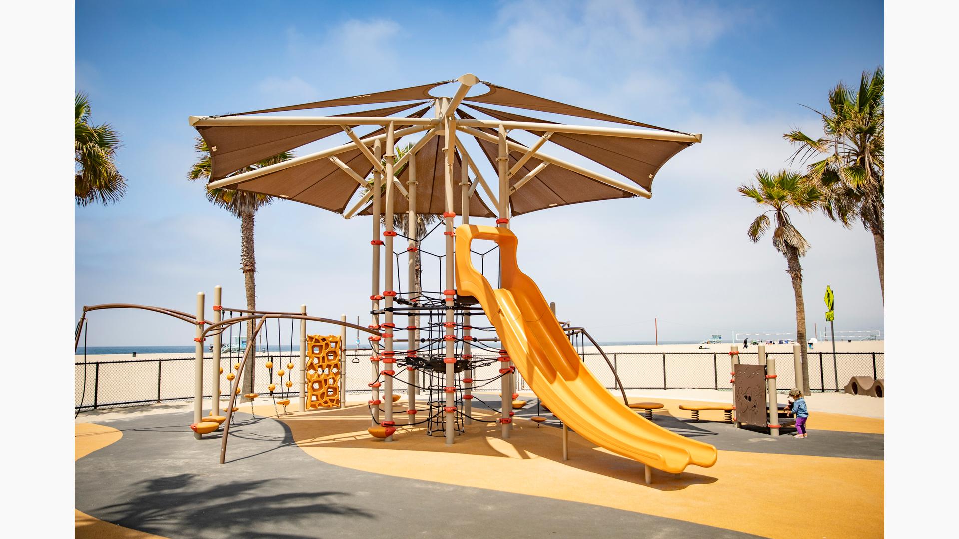 A large playground tower with overhead shade system has a labyrinth of ropes for climbing, a large double lane slide and other connecting play activities like monkey bars, spinners, and climbers. The play area is fenced in surrounded by scattered palm trees and a large sandy beach leading to the ocean.