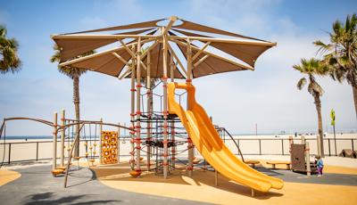 A large playground tower with overhead shade system has a labyrinth of ropes for climbing, a large double lane slide and other connecting play activities like monkey bars, spinners, and climbers. The play area is fenced in surrounded by scattered palm trees and a large sandy beach leading to the ocean.