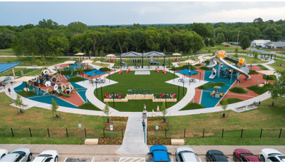 Full elevated view of a large park playground with a central artificial grass lounging area surrounded by play areas on either side with hexagonal shaped towers with slides and other hexagonal designed play structures.