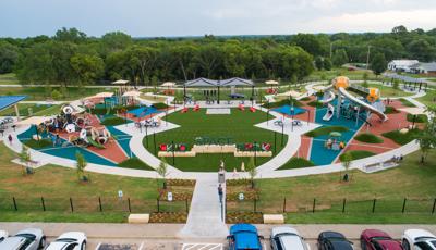 Full elevated view of a large park playground with a central artificial grass lounging area surrounded by play areas on either side with hexagonal shaped towers with slides and other hexagonal designed play structures.