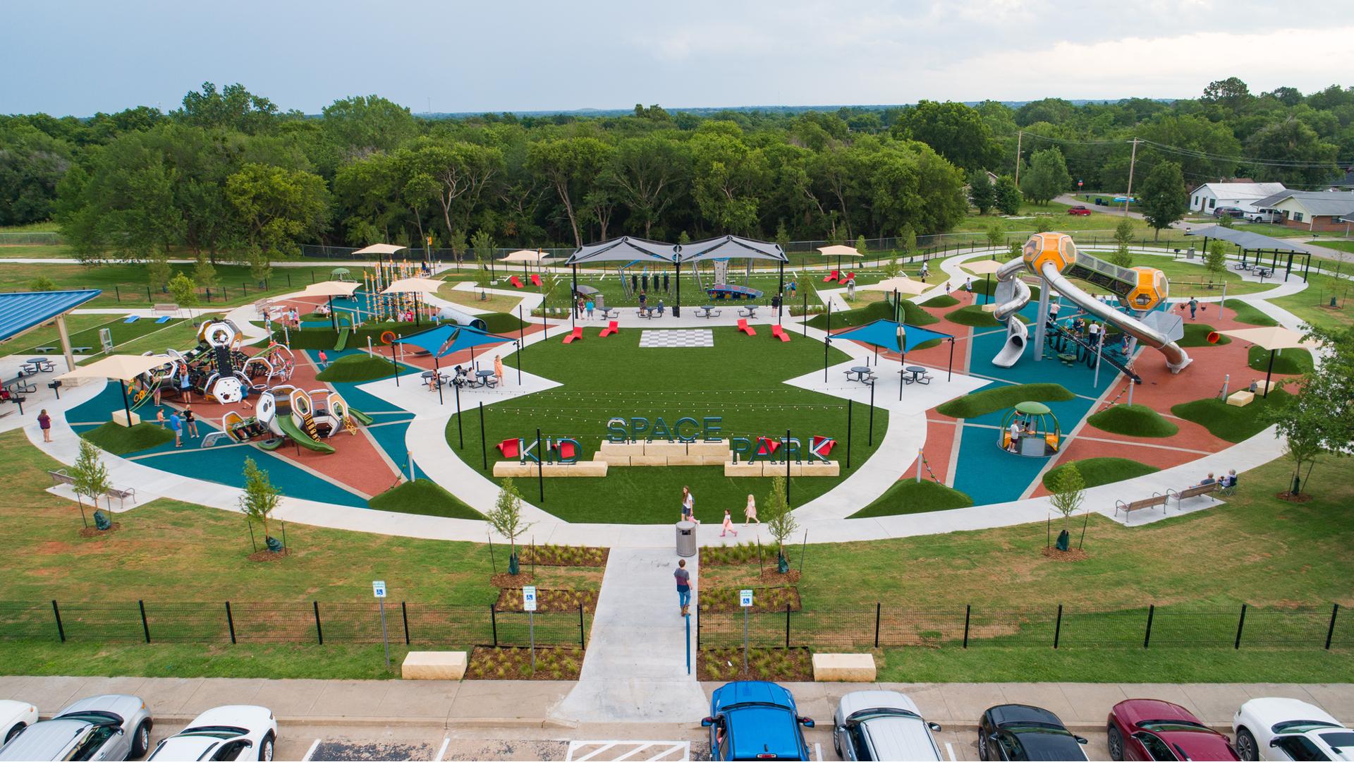 Full elevated view of a large park playground with a central artificial grass lounging area surrounded by play areas on either side with hexagonal shaped towers with slides and other hexagonal designed play structures.