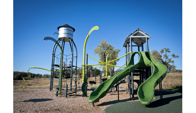 Green playground with tall net structure that has a silo type roof and tall posts shaped like wheat stalks. Double green slide in front with open grass and a few trees in the background.