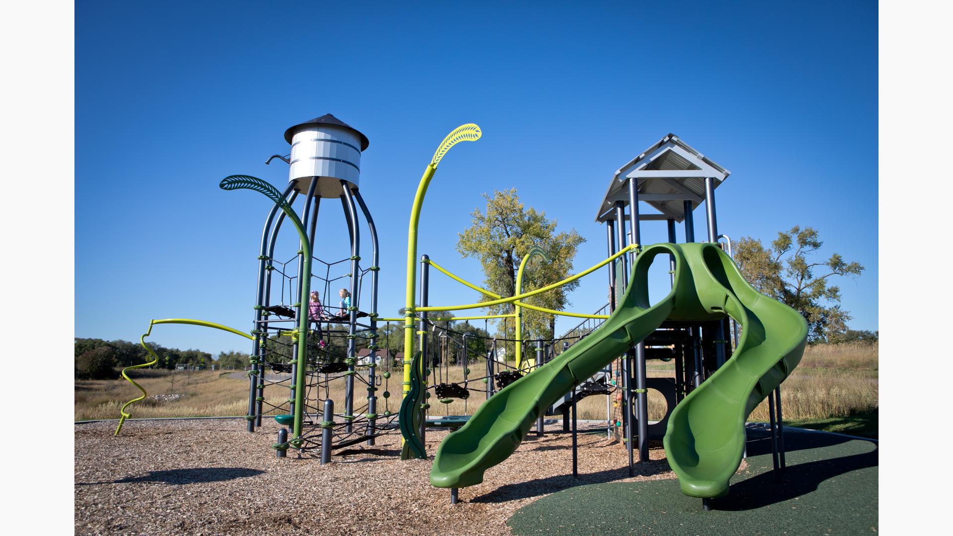 Green playground with tall net structure that has a silo type roof and tall posts shaped like wheat stalks. Double green slide in front with open grass and a few trees in the background.