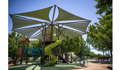 A play area surrounded by mature lush trees has a large tree fort like play structure tower with a large octagonal shade overhead. An additional arched ladder climber sits just to the right of the large play structure. 