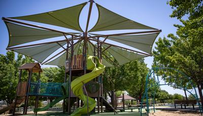 A play area surrounded by mature lush trees has a large tree fort like play structure tower with a large octagonal shade overhead. An additional arched ladder climber sits just to the right of the large play structure. 