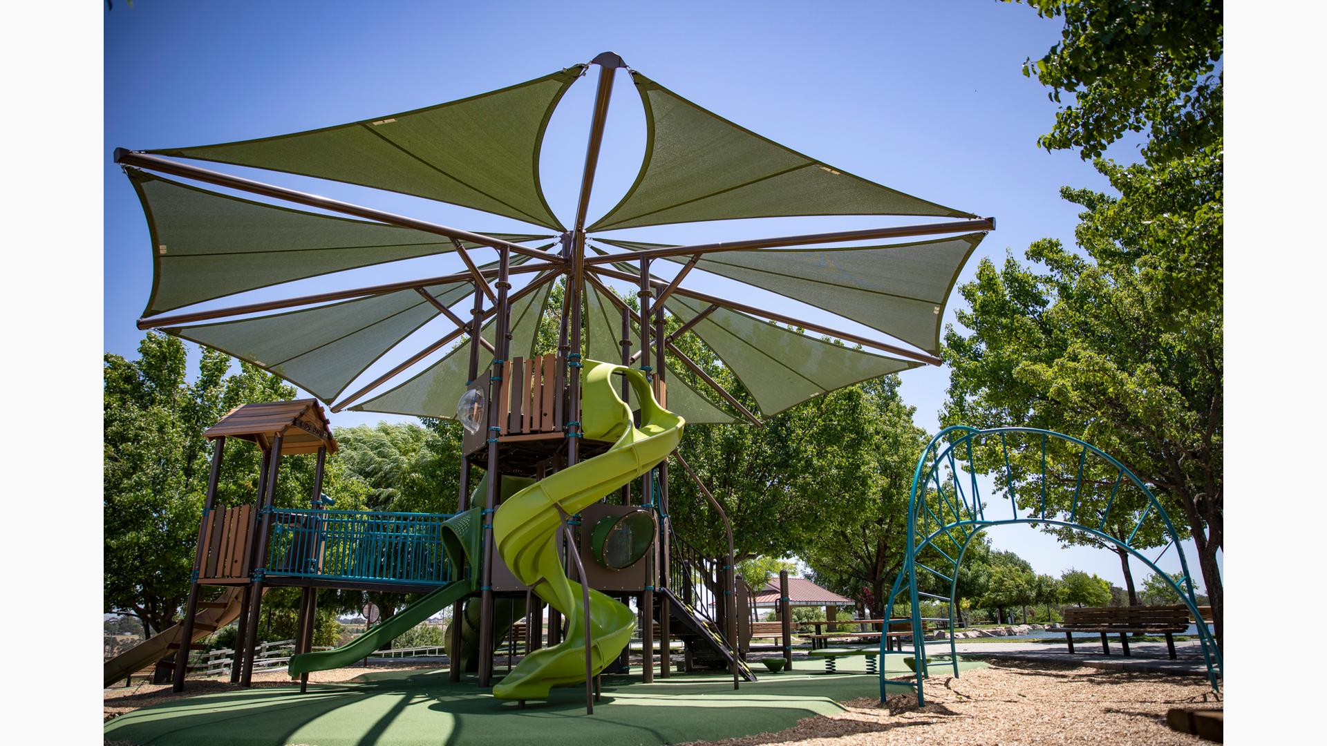 A play area surrounded by mature lush trees has a large tree fort like play structure tower with a large octagonal shade overhead. An additional arched ladder climber sits just to the right of the large play structure. 