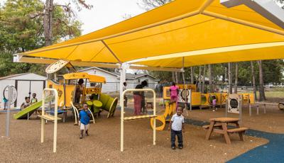 A large yellow shade covers a play area with two separate play structures for toddler aged children. Children and teachers play at the yellow and green colored play activities.