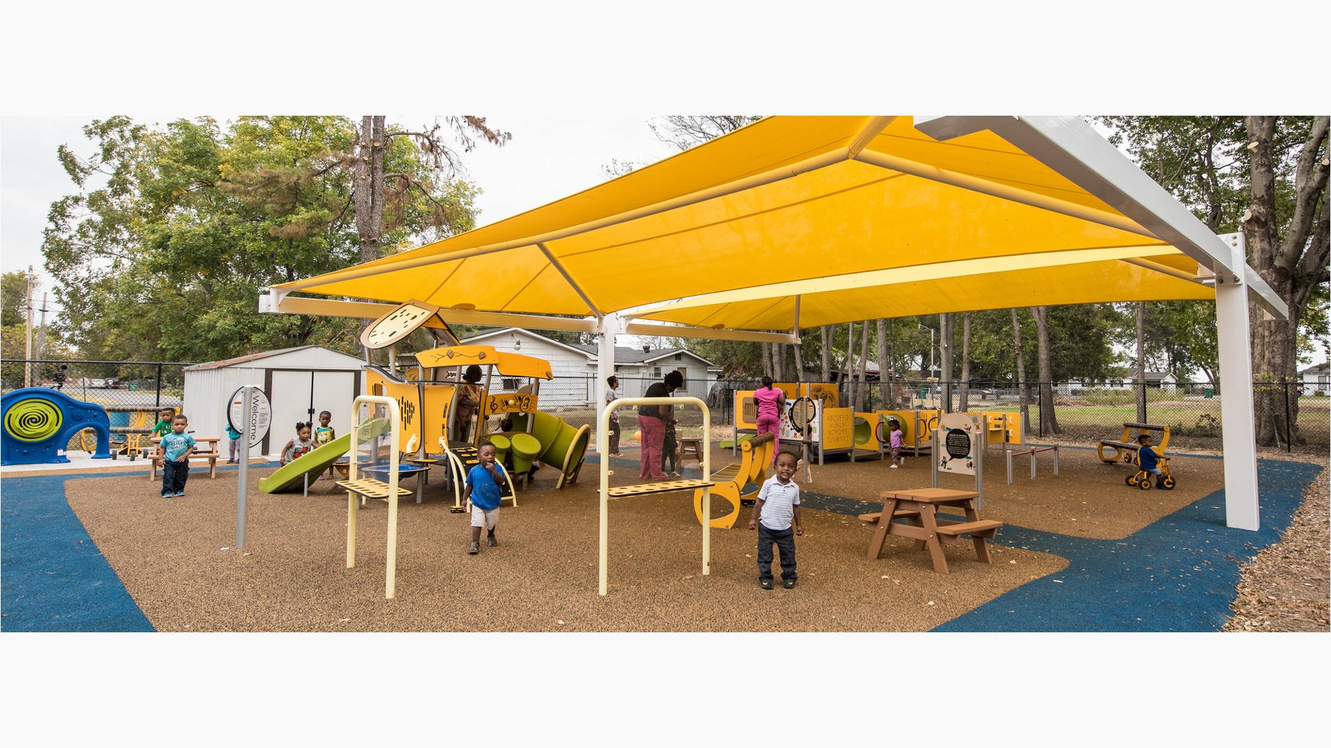 A large yellow shade covers a play area with two separate play structures for toddler aged children. Children and teachers play at the yellow and green colored play activities.